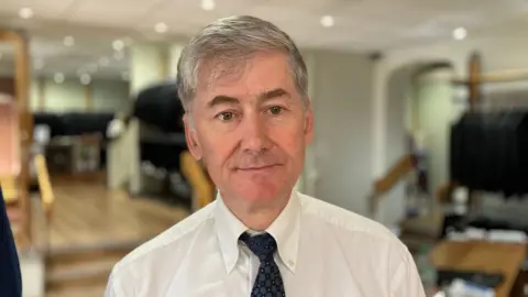 Neil Warwick, standing in a shop. He has grey hair and is wearing a white shirt, blue tie and a white shirt with an oxford collar.