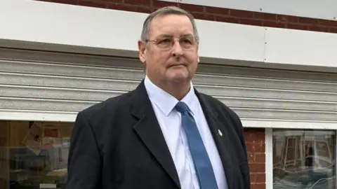 North Warkwickshire and Bedworth Conservatives A man with short grey hair, glasses, a black jacket, blue tie and white shirt stands in front of a building with the shutters partially down.