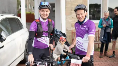 Family Picture On the right is a man stood next to a bike whilst smiling at the camera. He has dark eyes and is wearing a bicycle helmet on his head with sunglasses resting on the helmet. He has a purple t-shirt on with a black long sleeve top underneath and black shorts. Next to him is a man with grey hair who's also smiling at the camera. He's holding a bike too. He's wearing a purple t-shirt and black shorts.