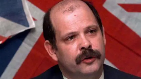 The late David Ervine, pictured from the shoulders up. He is balding and has dark brown hair and a brown moustache. there is a Union Jack flag on the wall behind him. 