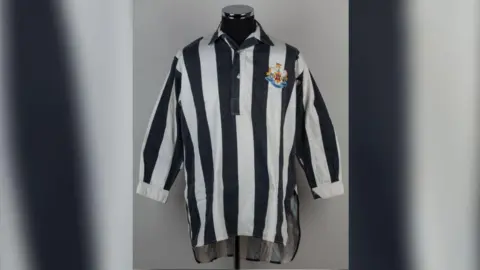 The 1952 black and white shirt Newcastle shirt displayed on mannequin. It has a Newcastle logo on the right hand side and long sleeves.