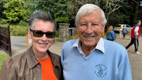 Lynn, a woman with grey with short grey hair and sunglasses with a brown jackey, she smiles next to Adrian a man with a light blue jumper and a white shirt with white hair, he also smiles.