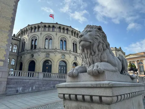 A large grey stone lion on a plinth, with an ornate round yellow building beyond flying the Norwegian flag from a pole on its roof