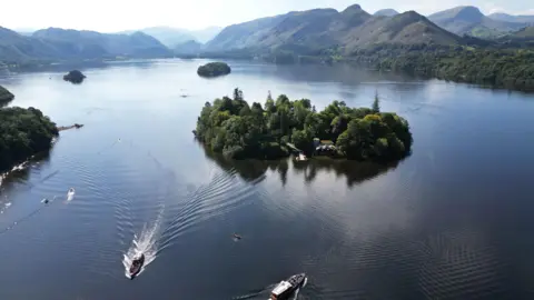 PA An aerial shot of Lake Windermere with boats going to and from the little island in the middle, which is covered in green foliage. The lake is ringed by large mountains.
