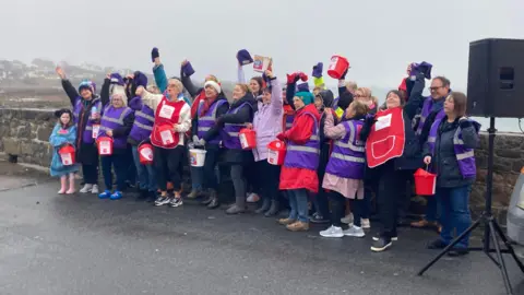 A group of people wearing purple vests hold red fundraising buckets up as the stand in front of a sea wall.
