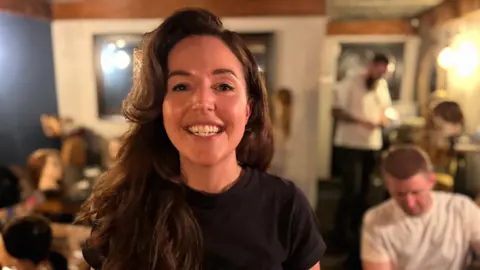 A woman with a side parting and long wavy brown hair looks at the camera smiling. She is wearing a black t-shirt. Behind her you can see dolls' heads and dads plaiting hair. 