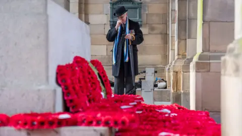 PA Media A veteran wearing a long, dark jacket with medals on his chest and a dark hat. He is carrying a dark umbrella with a wood-coloured handle. He is wiping his face while staring at a number of red wreaths laid on the floor next to a grey stone block. Behind him is a wall made of light brick. Light brick pillars can be seen to the right of the image.