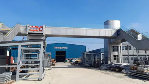 A footbridge at a depot site. It is made of stainless steel. A warehouse can be seen in the distance behind it. 