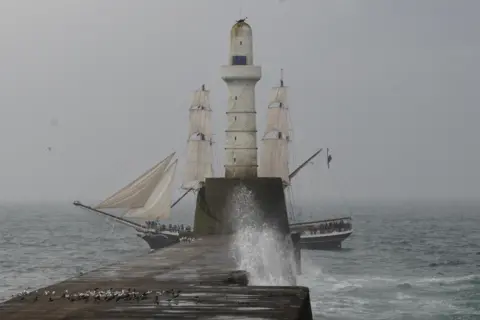 Sarah Sivers A ship sailing past a lighthouse on a stormy day with grey skies 