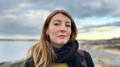 BBC A woman with long red hair wearing a black scarf and coat looks into the camera. behind him is the sea and a rock