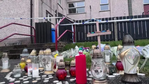 Candles and tributes on pavement outside a house. There are grass, steps and grey houses behind. 