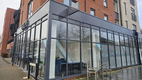 A black-framed glass shop front on the corner of a building with the word "CALM" in capital letters on a sign above and two chairs outside.