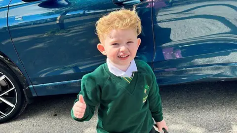 Kent Police Peter Maughan, a four-year-old boy with short, blond hair, is standing by a blue car wearing a school uniform. He has a white shirt, a green and yellow tie and a green jumper. He is holding a green schoolbag and is making a thumbs-up sign.