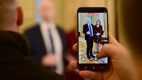 Photo of a hand holding an iphone, photographing Jon Burrows and Diana Armstrong