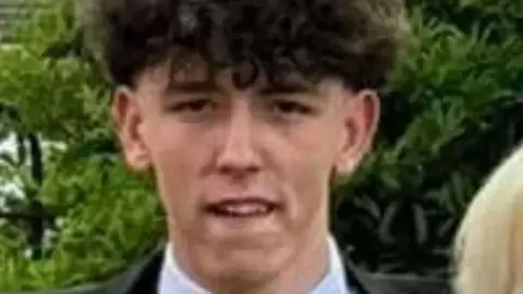 Bedfordshire Police Thomas Taylor, who has curly dark hair, is standing in front of green foliage. He is wearing a pale shirt and dark jacket.