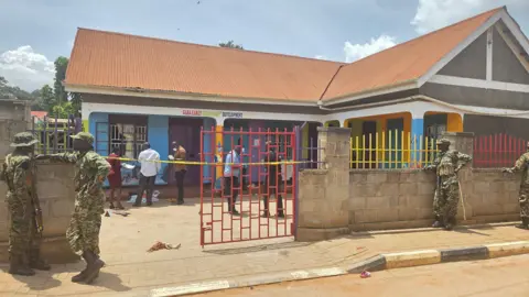 A low-rise school building with police tape around the gate. Men in military fatigues can be seen standing by the perimeter wall.