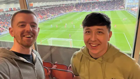 Darren Dowling (left) with short brown hair wearing a black t-shirt and grey hoodie and David Handling (right) with short brown hair and wearing a light green hooded top. They are standing in front of windows in a hotel room showing a pitch side view of Blackpool FC playing Bolton Wanderers with packed crowds. They are both smiling.
