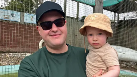 BBC News A man in a green jumper, sunglasses and a dark baseball cap holds a young child. The child is wearing a sand coloured top and bucket style hat. They are in front of an animal enclosure 