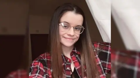 Zoe / Become A young woman with long hair and glasses, wearing a red checked shirt.