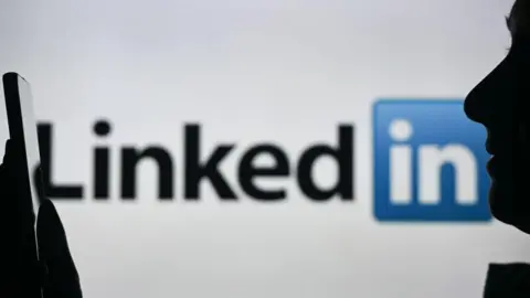 A silhouette of a woman holding a smartphone in front of a LinkedIn logo displayed on a computer screen behind her