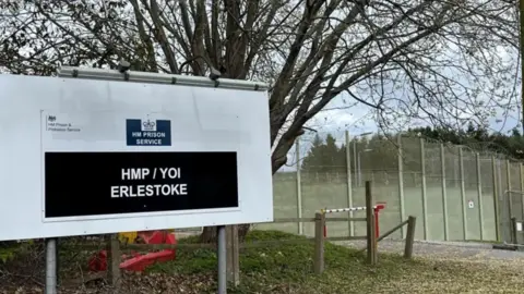 A sign which says 'HMP Erlestoke' with a fence behind it