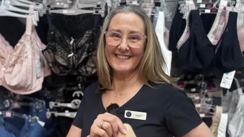 A woman wearing glasses stood smiling, wearing a uniform in a shop. 