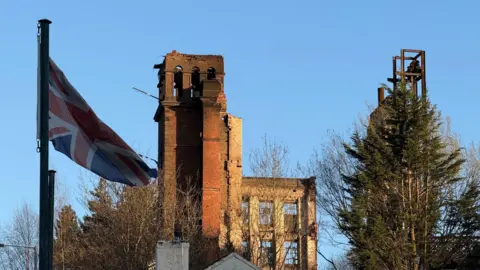 Part of a brick-built mill building. There is a union jack flag in the foreground and trees obscuring part of the structure. The scene is seat against a blue sky.