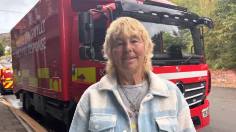 A woman with blonde hair looking at the camera. She has a blue and white woolly jumper. There is a red fire truck behind her.