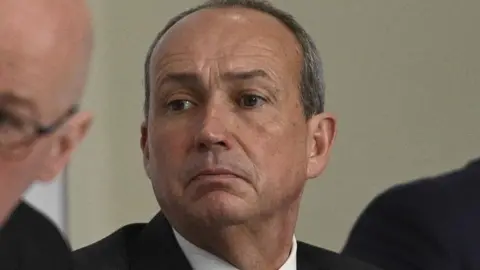 A close-up of Deputy Lyndon Farnham. He has short dark hair and is wearing a suit. He has a concerned expression.