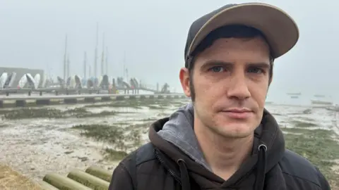 Peter Walker/BBC Justin Francis is a young man wearing a dark-coloured coat and hooded top, and a baseball cap. Behind him is the mud flats and boats of Brightlingsea Harbour on a foggy day.