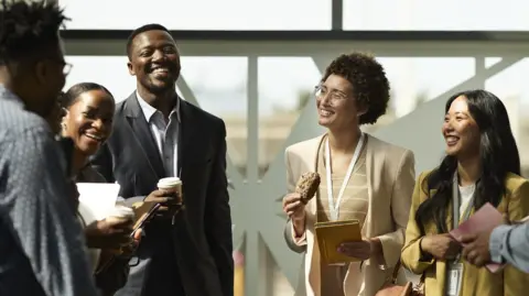Group of young people in professional clothes standing in a group smiling and laughing, holding coffees