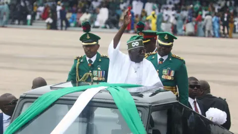 AFP via Getty Images Bola Tinubu in a white robe and green hat stands on the back of black vehicle decked in the green and white national colours of Nigeria. Behind him stand bemedaled military officers in green and black suited men in dark glasses surround the vehicle.