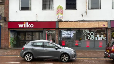 Martin Heath/BBC Shop front of two units. The one on the left has a Wilko logo on a red background above the entrance. The one on the right has the hoarding boarded up. The words "The Co Lab" are displayed in white lettering to the right. There is an "under offer" sign above the entrance and a grey car in front of the store.