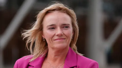 Sandy Peggie looking slightly off camera. She has fair hair and is wearing a bright pink blazer.