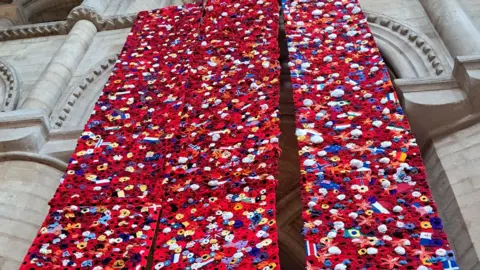 Peterborough Cathedral Three narrow, red hangings side-by-side. They are covered in handmade flowers. The hangings are suspended in Peterborough Cathedral. There are stone pillars behind them. 