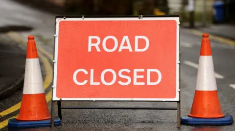 A red 'ROAD CLOSED' sign with two red and white cones on either side