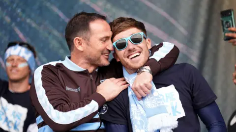 PA Media Frank Lampard, wearing a brown tracksuit top, hugs midfielder Josh Eccles.
