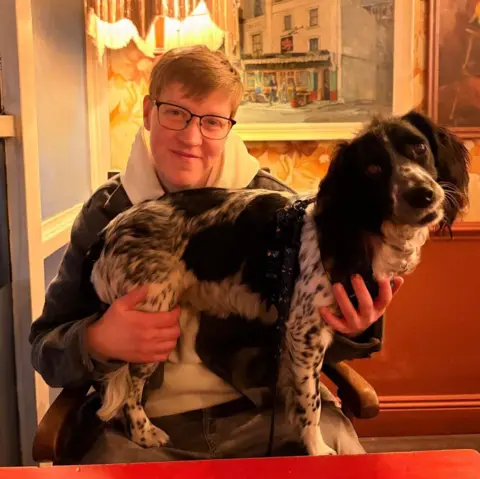 Supplied A woman sat at a table. She is smiling at the camera and she has a black and white spaniel dog standing on her knee. The dog is looking at the camera.