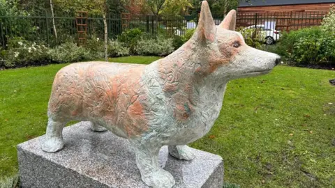 A sculpture of a corgi, made from cast metal, sits on a plinth by grass