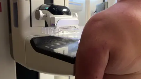 A close-up profile view of a patient standing at a mammogram machine during a breast screening appointment.