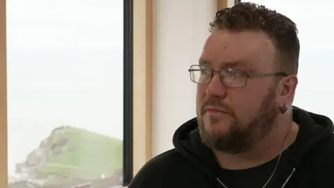 The image shows a person sitting indoors near a large window with a coastal view behind them. The person is wearing dark clothing, including a hoodie, and has short, curly hair.
