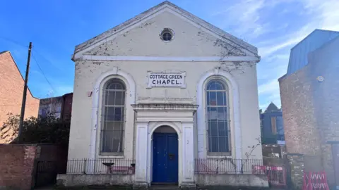 A cream coloured building with a blue door showing years of wear and tear with peeling paint and weeds growing from the cracks