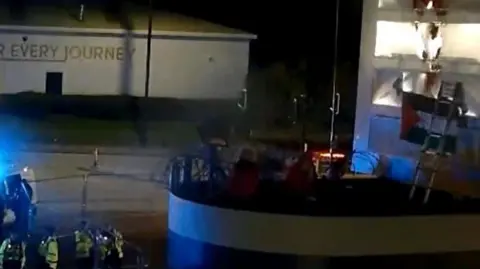CPS CCTV still of two women in red boiler suits sitting on top of a security building, with red paint splashed all over a large sign and a Palestinian flag hung up. Police officers in high vis coats are standing on the gorund looking up at them.