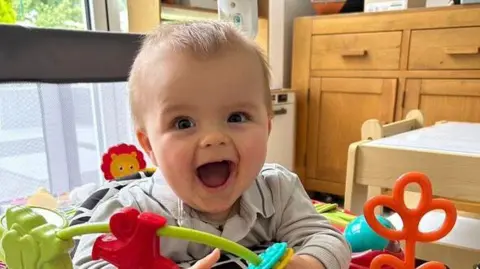 Mums Go Wild Swimming A baby, Kit Baddeley, is sitting in a high chair with toys surrounding him. He is looking into the camera and smiling.