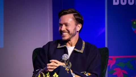 Wimbledon BookFest A man sitting on a chair while holding a microphone and smiling while looking towards the right. He is wearing a white shirt and a navy blue shirt over it with a embroidered collar. He is speaking at a panel with a purple background 