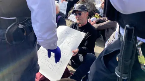 PA Del Naja sits on the floor near other fellow protestors as he holds a sign that reads: "I oppose genocide I support Palestine Action." Two police officers stand in front of him.