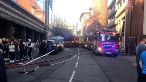 Four fire engines parked on the righthand side of a road, facing the camera and positioned next to a tall building which is the Crown Plaza Hotel. On the other side is a crowd of people.