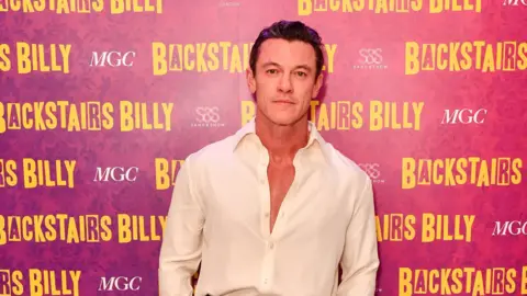 Getty Images Welsh actor Luke Evans standing in front of a background that says Backstairs Billy, he is wearing a whit shirt that is unbuttoned. 