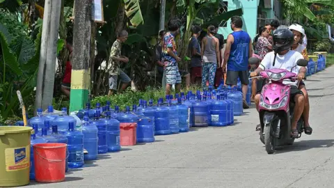 Getty Images Earthquake survivors lining up to refill blue containers of water as two people on a pink motorbike drive by