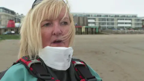 Kristen Cureton, with blonde hair and wearing a green waterproof jacket, and white face-mask speaks to the camera 